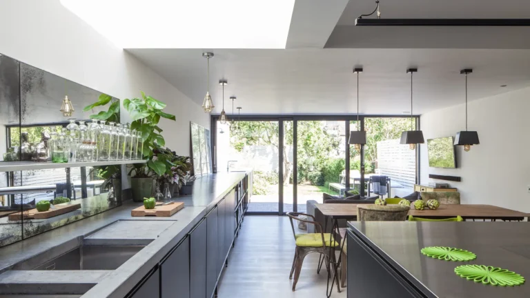 Modern kitchen and dining area in North London, designed by Trevor Brown Architect, features large windows, sleek cabinetry, and plants. Bright, natural light illuminates the room with a view of the outdoors.