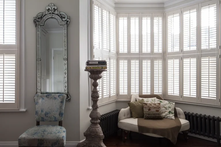 In a charming Harringay home, a room features a large mirror and a vintage chair. By the window with white shutters, a cozy sofa—part of the recent home refurbishment—rests adorned with pillows and a throw, while books grace the pedestal beside it, exemplifying North London elegance.