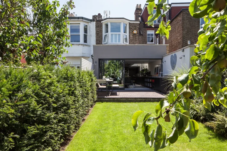 A two-story brick house features a modern extension by Trevor Brown Architect, viewed from the backyard. The garden, with its manicured lawn, hedges, and trees, leads to an open glass patio door.