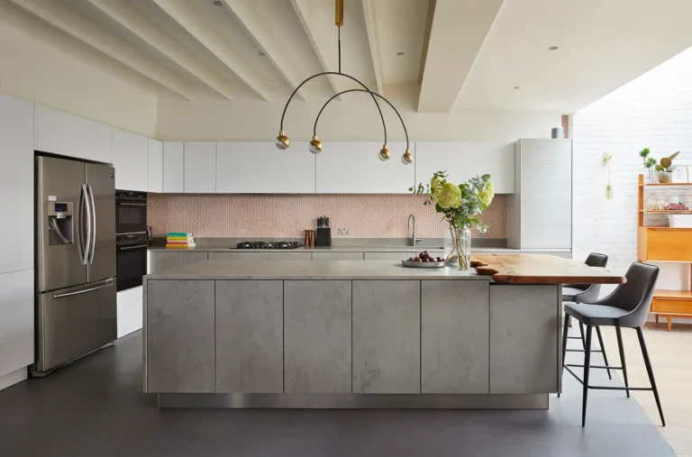 Modern kitchen in North London with a concrete island, built-in appliances, white cabinetry, and pendant lighting. As part of the home's refurbishment, two barstools are at the island, with a vase of flowers gracing its countertop—a touch of impeccable interior design.