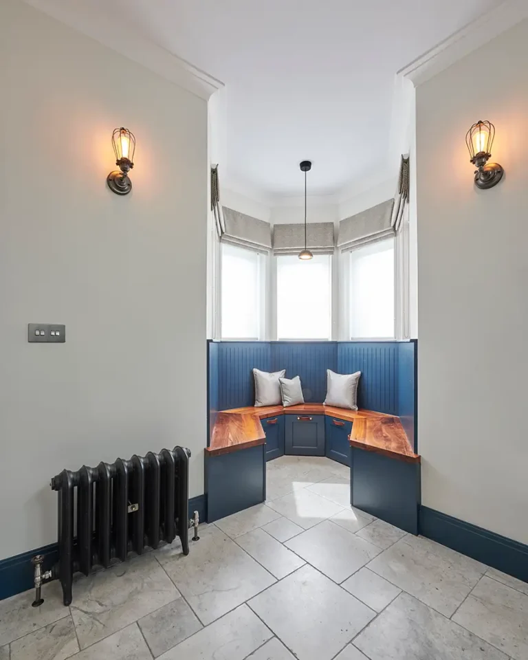 A cozy nook in Muswell Hill with a wooden bench and pillows, set below tall windows. Wall lights softly illuminate the space, capturing the essence of North London's charm, while a black radiator sits nearby on a tiled floor. Perfect for those who appreciate thoughtful interior design.