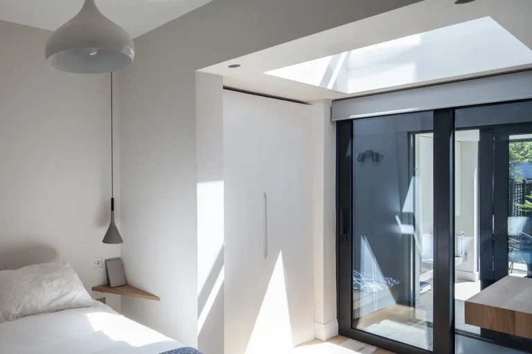 This small modern bedroom, recently enhanced through home refurbishment, features a skylight and sliding glass doors opening to a balcony. A pendant light elegantly hangs above the bedside table adorned with a book and pillow on the bed, embodying chic interior design in Bounds Green.