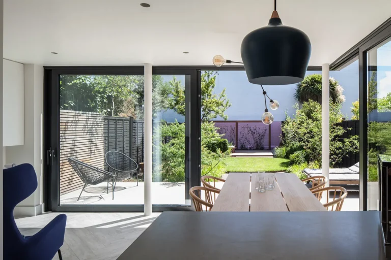 A modern dining area, designed by Trevor Brown Architect, features a large wooden table surrounded by chairs and overlooks a lush garden through floor-to-ceiling glass doors. A black pendant light hangs above the table in this elegant North London extension.