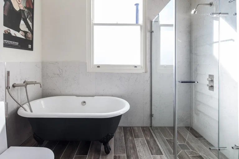 A bathroom in North London features a clawfoot bathtub, a glass-enclosed shower, and a window. Designed by Trevor Brown Architect, the space is completed with a poster on the wall and tiled with brown wood-like tiles for an elegant interior design touch.