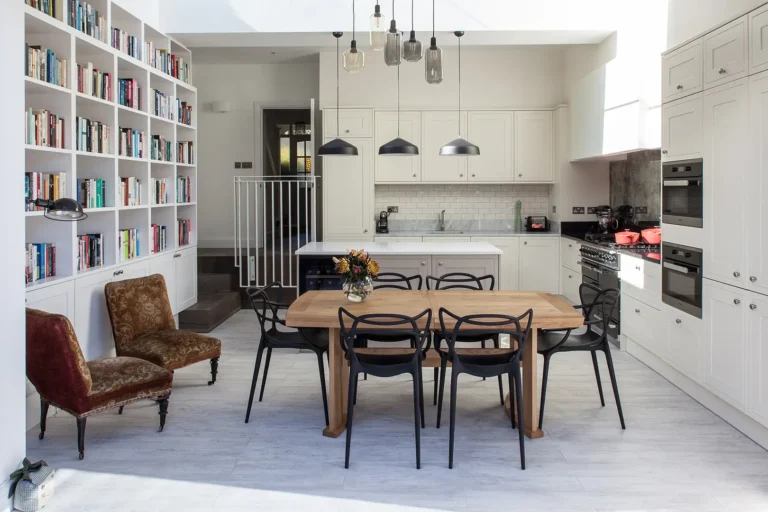 Designed by Trevor Brown Architect, this North London kitchen extension features white cabinets, a wooden dining table, black chairs, and pendant lights. A cozy nook with a bookshelf and two armchairs sits to the left.