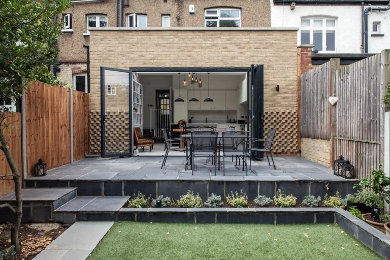 A modern patio designed by Trevor Brown Architect features a dining set, large glass doors leading to the kitchen, and is enveloped by wooden fences and lush greenery.