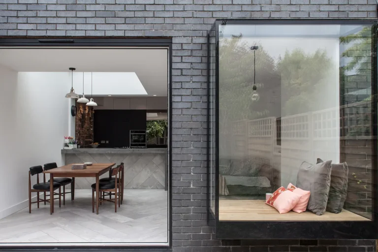 Modern home with a dining area visible through a large window, designed by Trevor Brown Architect. It features a wooden table and chairs. Adjacent is an extension housing a cozy window nook with cushions, set against a dark brick exterior reminiscent of Alexandra Palace's charm.