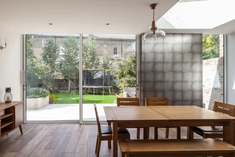 Bright dining room designed by Trevor Brown Architect in North London, featuring a wooden table and chairs. Large sliding glass doors open to a garden with a trampoline, while a skylight above and textured wall on the side enhance the Islington charm.