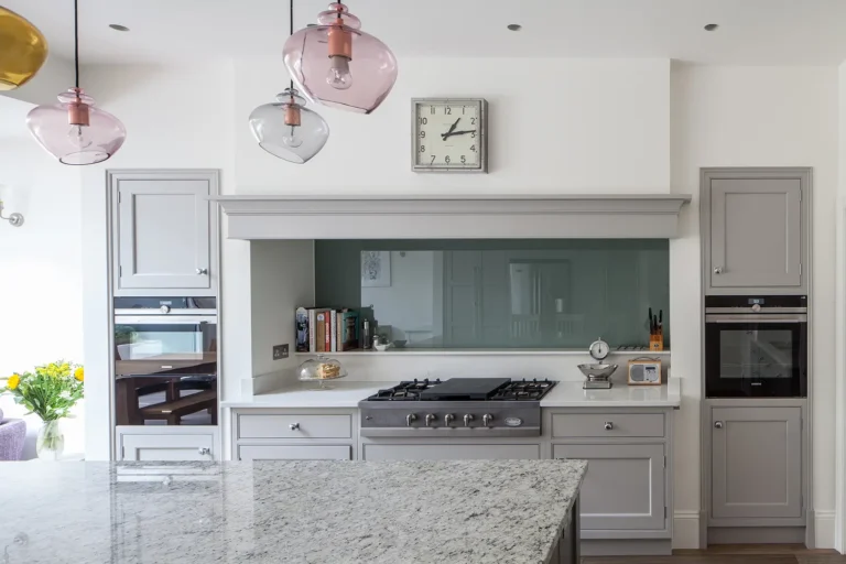 A modern kitchen in North London boasts gray cabinets, a large granite island, and stainless steel appliances. As part of the home refurbishment, pink pendant lights illuminate the space, while a wall clock and books add charm above the stovetop.