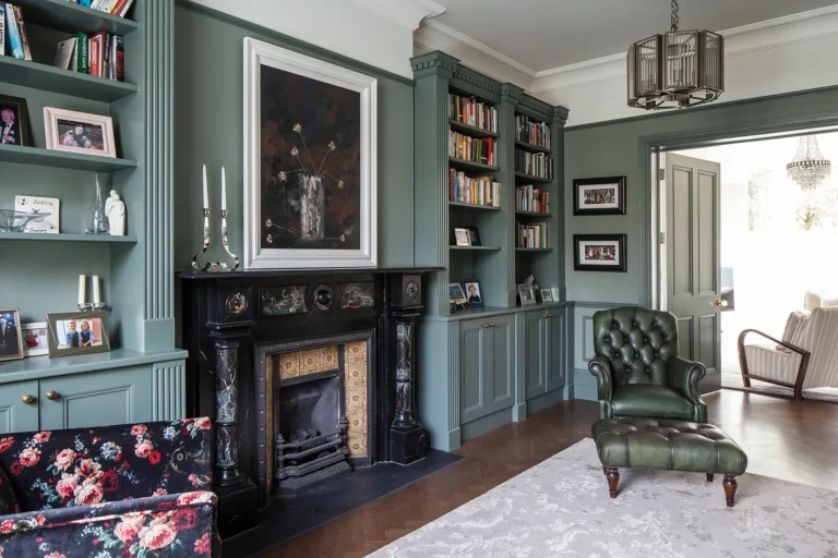 A cozy study featuring green walls and built-in bookshelves, crafted with the elegant touch of Trevor Brown Architect. The space includes a black fireplace, a green leather armchair, a floral couch, and framed photographs that enhance its charm.