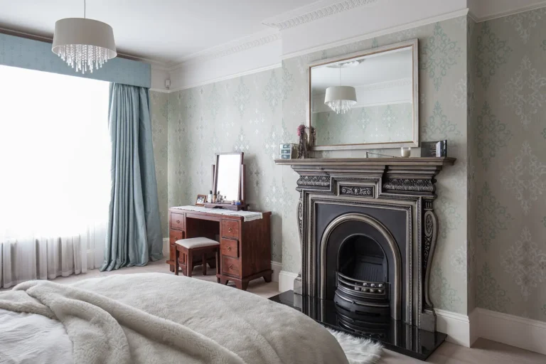 Elegant bedroom in a North London extension, designed by Trevor Brown Architect. Featuring a vintage fireplace and an ornate mirror above, complemented by a wooden dressing table. Pale patterned wallpaper and large windows with teal curtains complete the décor.