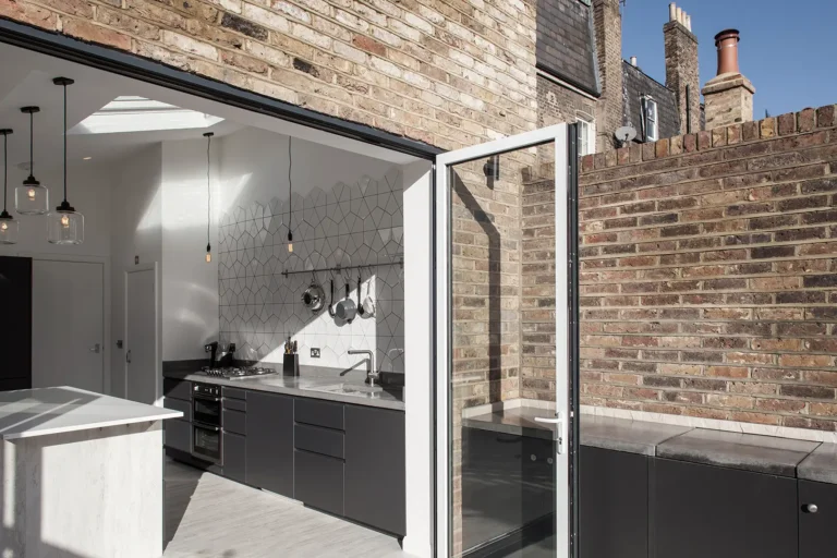 Modern kitchen designed by Trevor Brown Architect in Hackney, featuring dark cabinets, marble countertops, and a geometric tile backsplash. An open glass door leads to a charming brick-walled outdoor area. Pendant lights hang elegantly from the ceiling, completing this home refurbishment.