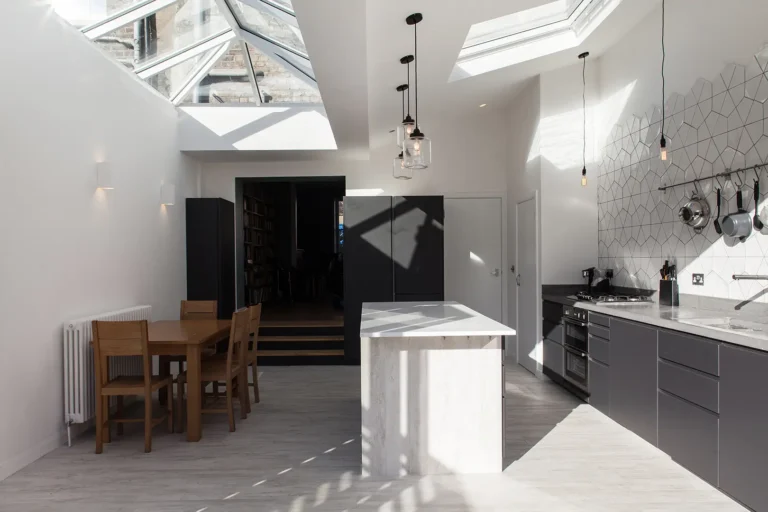 A modern kitchen in Hackney features a central island, skylights, and geometric wall tiles. The stylish interior design includes a wooden dining table with chairs on the left, while elegant pendant lights hang from the ceiling as part of the home's recent extension.