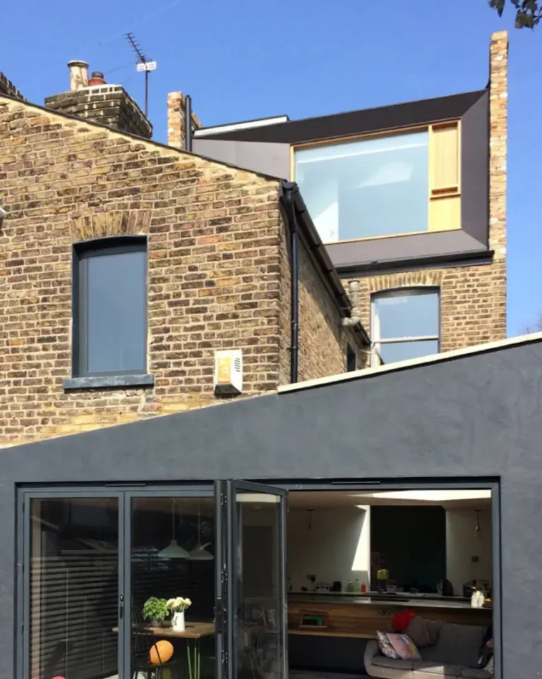 This North London home refurbishment showcases a modern extension on an old brick building, featuring large glass doors that open to a contemporary interior and an upper loft with a grand window.