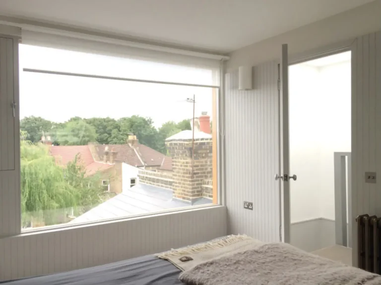 A bedroom designed by Trevor Brown Architect features a large window overlooking rooftops and trees. This interior design includes a bed, a radiator, and a partially open door leading to the bright extension beyond.