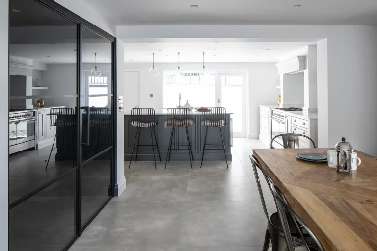 Modern kitchen designed by Trevor Brown Architect features a central island, black bar stools, and white cabinetry. A large wooden dining table sits beneath pendant lights, while a glass partition completes the home's stylish refurbishment.