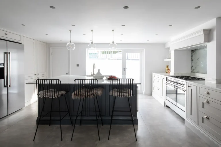 Designed by Trevor Brown Architect, this modern Tufnell Park kitchen features a large island with three bar stools, stainless steel appliances, and pendant lighting. White cabinetry and light gray accents create a clean, minimalist look in this stylish extension.