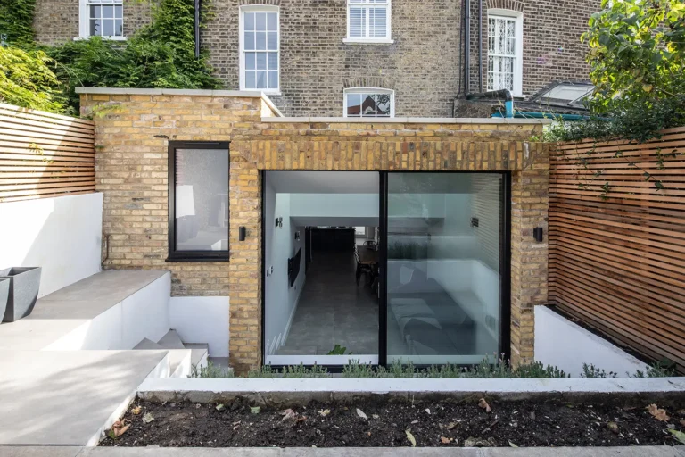 Modern brick house extension in Tufnell Park with large glass sliding doors, nestled into a landscaped garden featuring wooden slat fencing and a small patio. This North London home refurbishment seamlessly blends style and functionality.