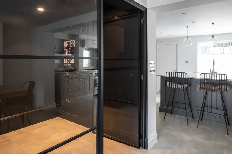 Modern kitchen in North London with a minimalist design, featuring a gray island and high chairs. Pendant lights illuminate the space, while a reflective glass partition provides subtle separation. Text on the wall reads "AFTER THE DISCO," adding a touch of flair to this Tufnell Park interior design marvel.