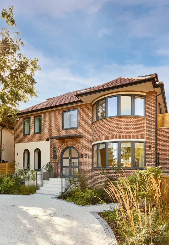 Trevor Brown Architect — Two-storey brick house with large bay windows, arched doorway, and landscaped front garden under a blue sky, designed by Trevor Brown Architect.