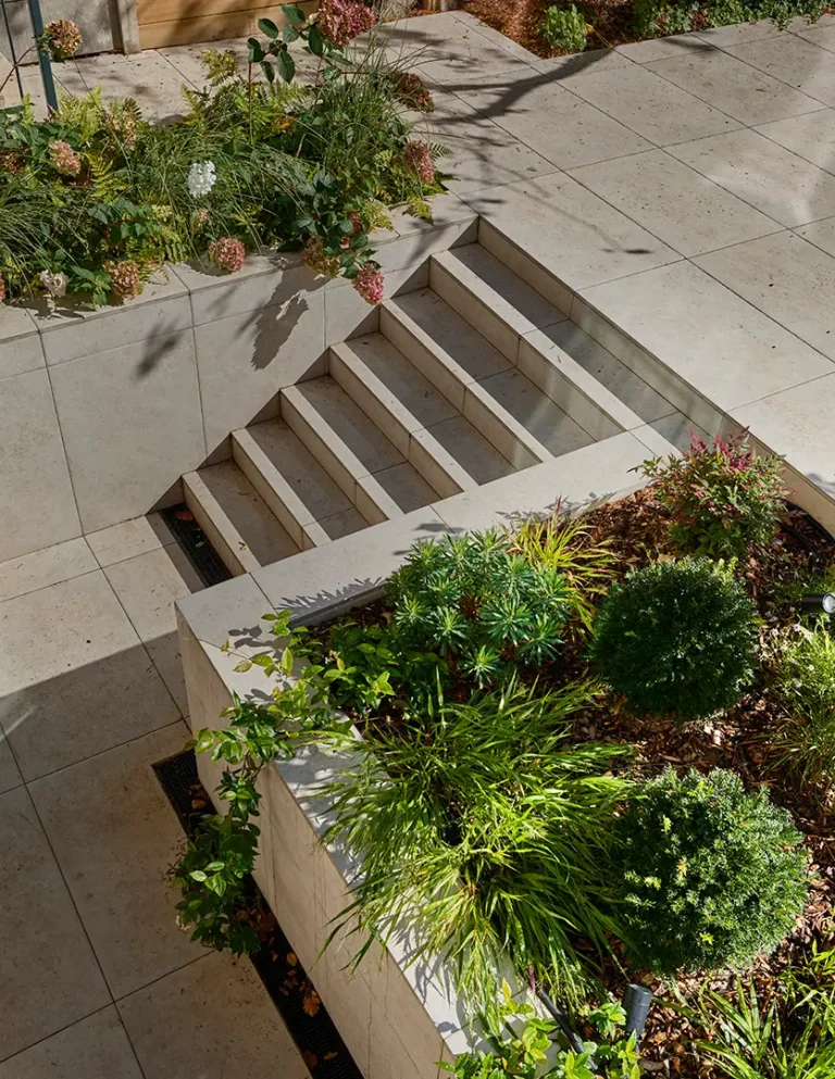 Stone steps lead down between two raised garden beds with green shrubs and plants, surrounded by large stone tiles in a landscaped outdoor area designed by Trevor Brown Architect.