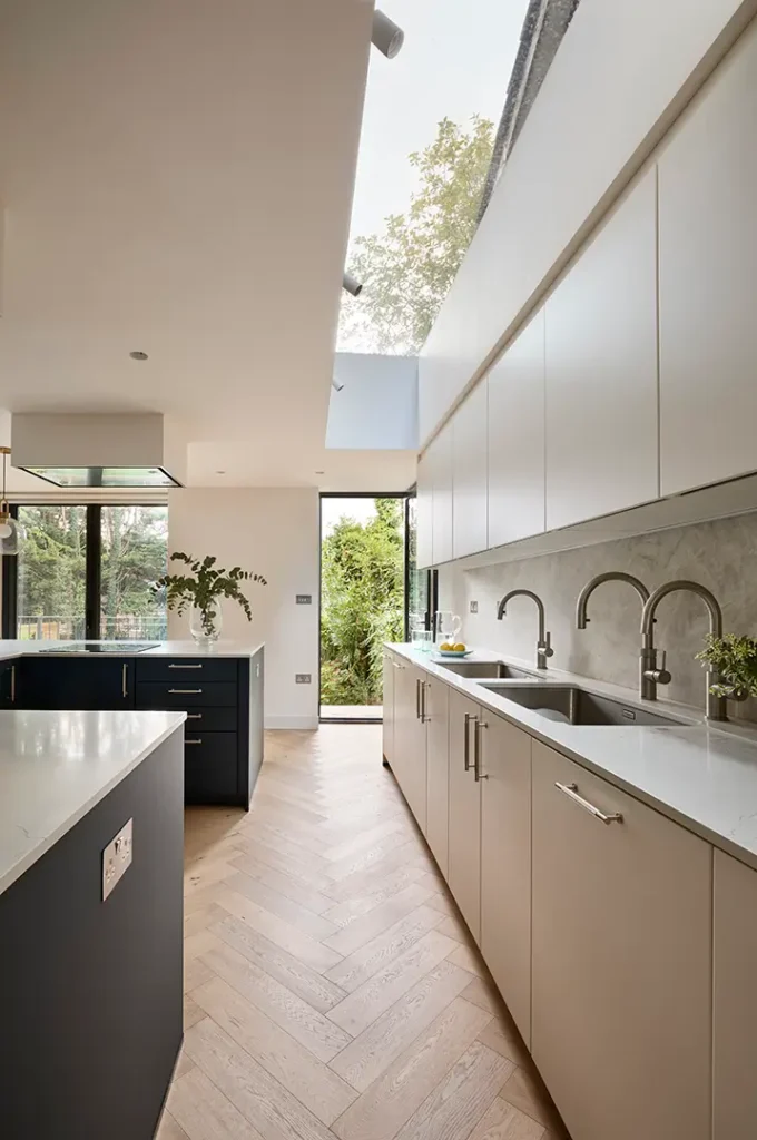 Trevor Brown Architect — Modern galley kitchen by Trevor Brown Architect with light wood floors, white cupboards, double sinks, marble splashback, skylight, and large windows
