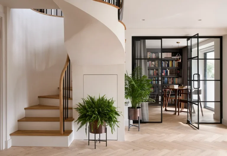 A modern interior by Trevor Brown Architect features a spiral wooden staircase, potted plants, and a glass door leading to a book-filled office with a large window.