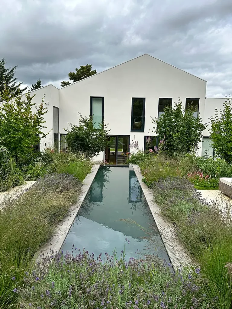 Modern white house with tall narrow windows and a rectangular reflecting pool, surrounded by lush, overgrown garden plants—an inspiring example of Trevor Brown Architect’s approach to home refurbishment and interior design extension.