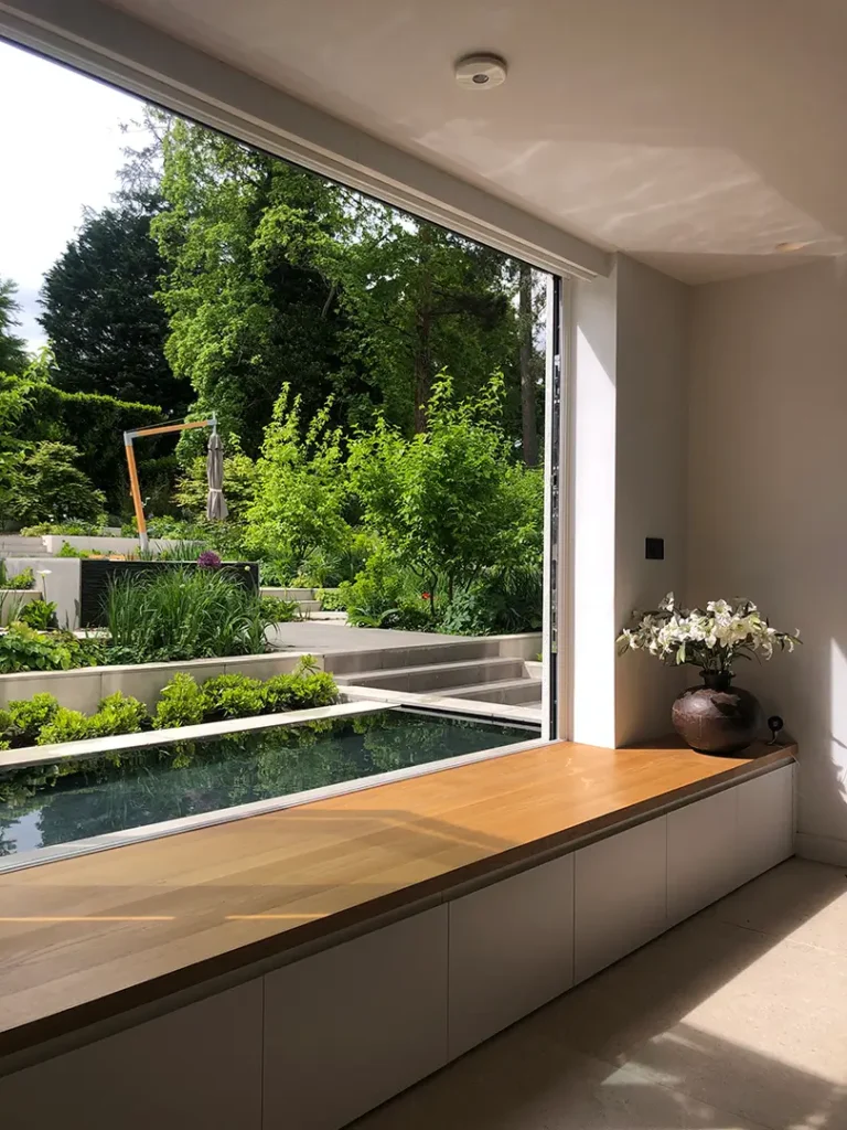A large open window reveals a modern garden with terraced landscaping, a pond, and lush greenery, viewed from a minimal indoor seating area with a vase of white flowers—an elegant showcase of Trevor Brown Architect’s interior design extension expertise.